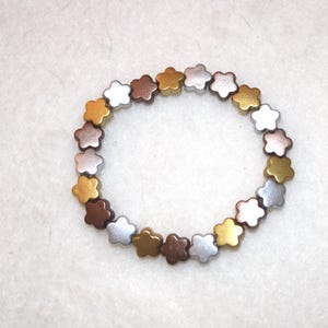 Könnte beinhalten: Ein Armband mit blütenförmigen Perlen in Gold-, Silber- und Bronzetönen. Die Perlen sind zu einem kreisförmigen Armband aufgereiht. Die Perlen sind gleichmäßig verteilt und bilden ein dekoratives Accessoire. Umfang: ca. 18 cm.