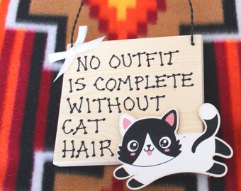 May include: A wooden sign with the text "No outfit is complete without cat hair" and a cartoon black and white cat with a pink nose and tongue sticking out.