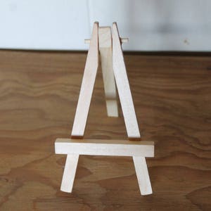 Może przedstawiać: Mały drewniany sztaluga, idealny do wyświetlania małych dzieł sztuki lub zdjęć. Sztaluga jest wykonana z naturalnego drewna i ma prosty, klasyczny design.