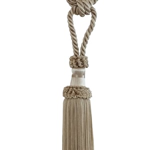 May include: A beige tassel tieback with a knotted rope design. The tassel is made of a soft, woven fabric and has a fringe at the bottom.