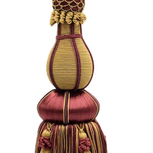 May include: Decorative tassel in gold, burgundy, and purple. The tassel has a twisted rope loop, a bulbous body, and multiple tiers of fringed tassels. A stylish accent for home decor.
