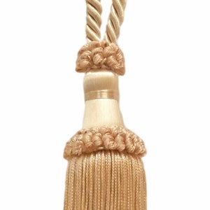May include: A beige tassel tieback with a knotted rope design. The tassel is made of a soft, woven fabric and has a wooden bead at the top.
