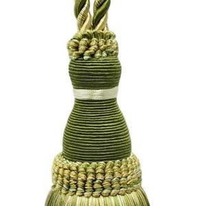 Puede incluir: Borla decorativa verde oliva y dorada con lazo superior. La borla presenta un lazo de cordón trenzado, un cuerpo cilíndrico y un fondo con flecos. Una banda plateada adorna el cuerpo.