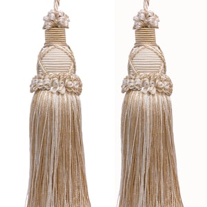 May include: Two decorative tassels with a beige and cream color scheme. Each tassel features a looped top, a detailed central section, and a long, flowing fringe. The tassels are designed for home decor, such as curtains or pillows.
