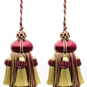 Pode incluir: Duas borlas decorativas em bordô e dourado. Cada uma apresenta um topo arredondado bordô com detalhes dourados, um corpo listrado em ouro e bordô e uma franja de borla dourada. Penduram de um cordão torcido, ideal para decoração de interiores.
