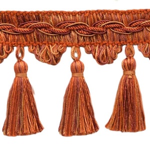May include: Decorative fringe trim in shades of orange and brown. The trim features a woven top section with a scalloped edge and hanging tassels. The tassels are a darker shade of orange, adding depth to the design. This trim is suitable for home decor projects.