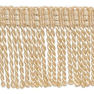 May include: A close-up of a beige fringe trim with a woven texture. The fringe is made of twisted threads and is approximately 1 inch in length.