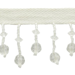 May include: Cream-colored beaded fringe trim. The trim features a woven fabric base with a chevron pattern. Clear, faceted beads hang from the base, adding a decorative element. This trim is suitable for various craft and design projects.