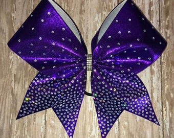 May include: Purple cheer bow with silver glitter and rhinestones. The bow is tied with a black elastic band.