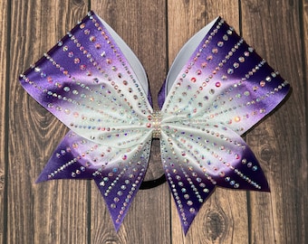 May include: A purple and white ombre cheer bow with rhinestones. The bow is tied with a black elastic band.