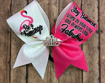 May include: White and pink glitter cheer bow with a pink flamingo and the text "Be a Flamingo" on one side and the text "Stay Balanced Stand by your flock and always look Fabulous" on the other side. The bow has a silver crown in the center.