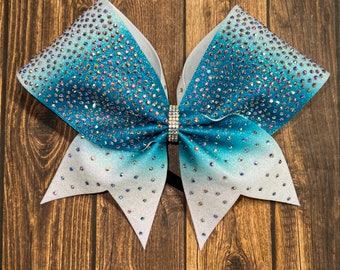 May include: A large white and teal cheer bow with iridescent glitter and rhinestones. The bow is tied with a black ribbon.