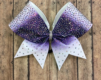 May include: A large white cheer bow with a purple ombre glitter design and clear rhinestones. The bow is tied with a black elastic band.