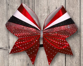 May include: A red, white, and black cheer bow with rhinestones. The bow is made of shiny fabric and has a black elastic band.
