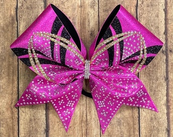 May include: A pink cheer bow with black and gold glitter accents. The bow is decorated with rhinestones and has a black and gold ribbon.