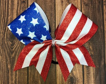 May include: A red, white, and blue glitter cheer bow with a star-spangled design. The bow is tied with a black ribbon.