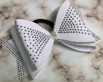 May include: White glitter hair bow with a large, sparkly rhinestone design. The bow is attached to a black elastic hair tie.