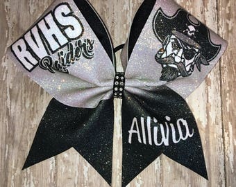 May include: A black and silver glitter cheer bow with a pirate logo and the text "RVHS Raiders" on one side and the name "Alliwia" on the other side.
