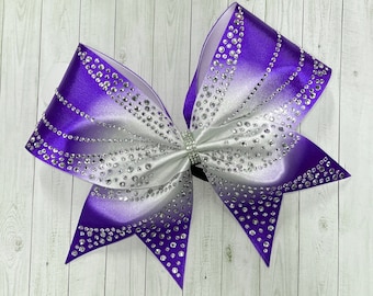 Pode incluir: Um grande laço de torcida roxo e branco com glitter prata. O laço é feito de fita de cetim e tem um design degradê.