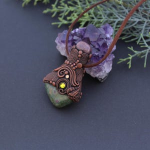 Può includere: Un ciondolo in pietra verde con un design in filo di rame e una piccola pietra preziosa gialla. Il ciondolo è appeso a un cordino in pelle marrone.