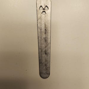 Puede incluir: Colgante de metal plateado con un símbolo de radiación recortado. El colgante tiene forma rectangular alargada con un extremo redondeado y un agujero circular en la parte superior para una cadena. El símbolo de radiación está en el centro.