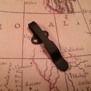 May include: A black metal pocket clip for a knife or multi-tool. The clip is designed to attach to a belt or pocket.