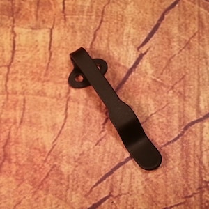 May include: Black metal pocket clip with a rounded tip and two holes. The clip is designed to attach to a pocket or belt.
