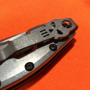 May include: Close-up of a silver pocket knife with a skull design on the pocket clip. The knife has a metal handle and black screws. The background is a bright orange fabric.