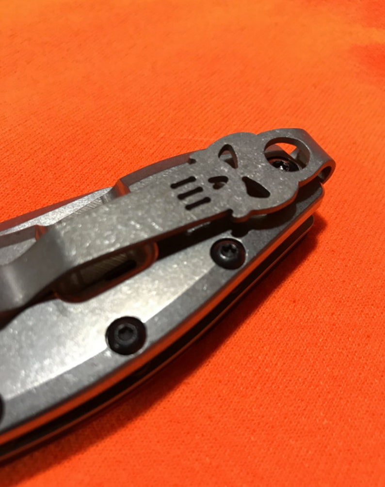 May include: Close-up of a silver pocket knife with a skull design on the pocket clip. The knife has a metal handle and black screws. The background is a bright orange fabric.
