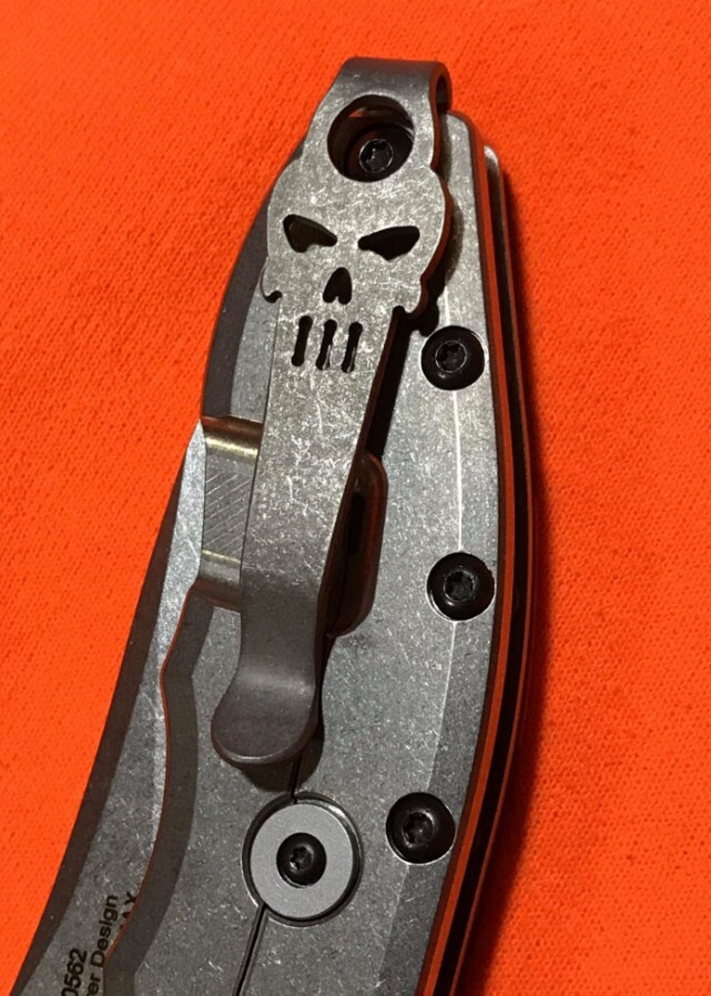 May include: Close-up of a silver folding knife with a skull-shaped pocket clip. The knife has a textured metal handle and black screws. The blade is not visible. The background is a solid orange color.
