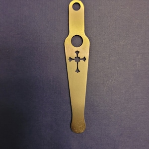 May include: A gold metal bottle opener with a cross-shaped cutout. The opener has a hole at the top for hanging.