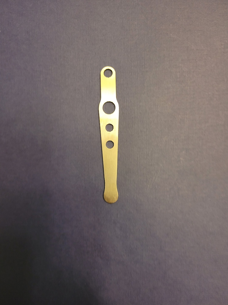 May include: A brass-colored metal tool with a flat, elongated design. It features a series of circular holes of varying sizes along its length, with a small hole at the top and a rounded end. The tool is set against a dark background.