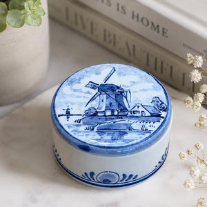 May include: A round ceramic trinket box featuring a blue and white hand-painted scene of a traditional Dutch windmill on the lid. The sides of the container are decorated with blue floral motifs on a white background, creating a classic aesthetic.