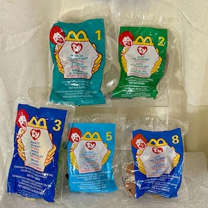 May include: Five Ty Beanie Babies in their original McDonald's Happy Meal packaging. The toys are:  Pickles the Leopard, Antsy the Anteater, Moochy the Frog, Rocket the Blue Jay, and Nuts the Squirrel. Each package has a number on it, and the Ty logo is visible.