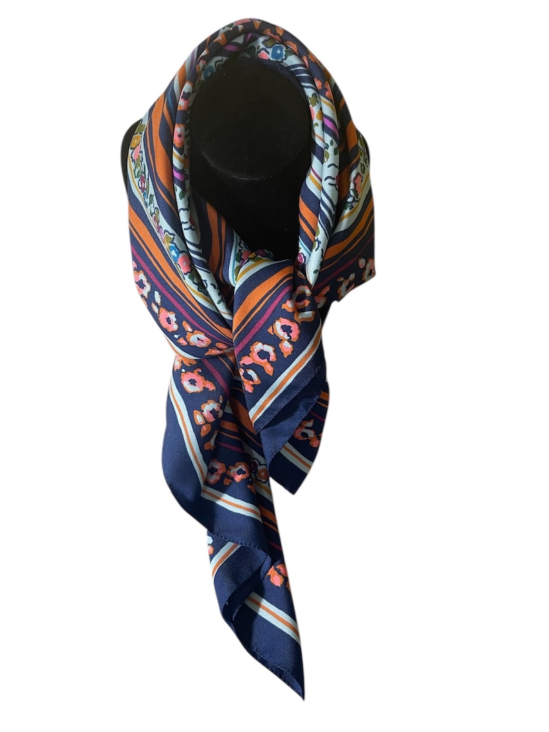 May include: A navy blue silk scarf with a floral and striped pattern. The scarf features orange, light blue, and pink floral accents. The scarf is draped over a black mannequin head.