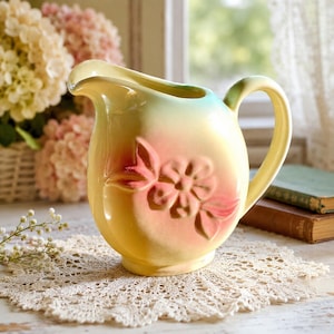 May include: A ceramic pitcher featuring a rounded body with a pale yellow and soft green gradient finish. The front displays a raised, embossed floral design in a muted pink hue. It has a curved handle and a wide, shaped pouring spout.