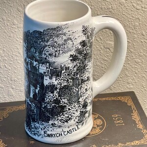 May include: White ceramic beer stein with a black and white illustration of a castle and trees. The stein has a handle and the words "Gwyrch Castle" printed on the bottom.