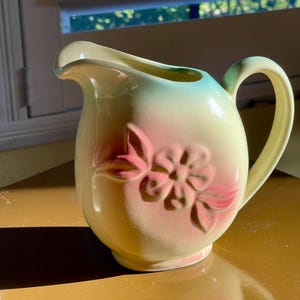 May include: A vintage ceramic pitcher with a floral design. The pitcher is yellow with a pink flower and green leaves. It has a light blue glaze on the top.