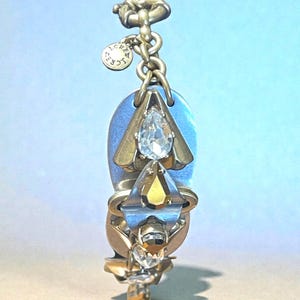 May include: A gold-toned pendant with a blue oval backing, adorned with clear and yellow crystal accents. The pendant has a teardrop-shaped crystal at the top and a diamond-shaped crystal below. A small circular charm with text hangs from the chain.