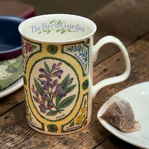 May include: White ceramic mug with a handle, featuring botanical illustrations and the text "The Herb Garden." The mug showcases images of herbs like sage, rosemary, and thyme, with a yellow border. A tea bag rests on a white saucer.