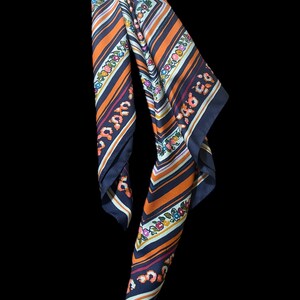 May include: A patterned scarf with a navy blue border, orange and white stripes, and floral accents. The scarf has a repeating floral pattern in pink, blue, and yellow. The scarf is made of a lightweight material.