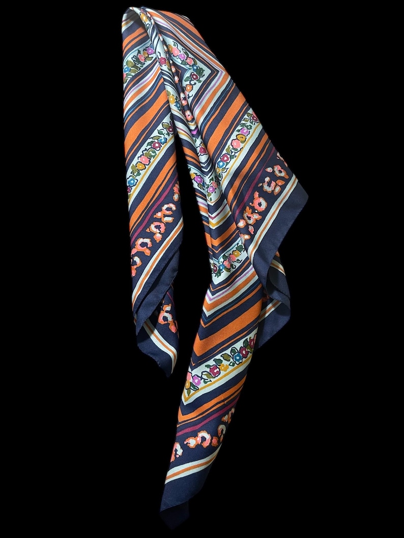 May include: A patterned scarf with a navy blue border, orange and white stripes, and floral accents. The scarf has a repeating floral pattern in pink, blue, and yellow. The scarf is made of a lightweight material.