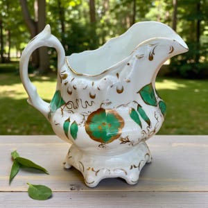 May include: White porcelain creamer with a scalloped rim, adorned with green floral and gold accents. The creamer has a decorative handle and sits on a pedestal base with three feet. A classic piece for serving cream or milk.