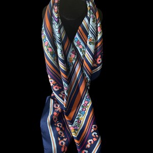 May include: A navy blue, orange, and white striped scarf with floral accents. The scarf is draped over a black form, showcasing its design. The scarf's edges are a solid navy blue.