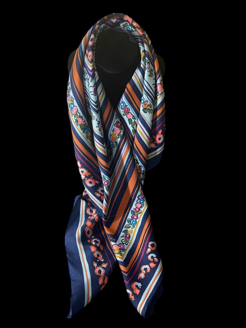 May include: A navy blue, orange, and white striped scarf with floral accents. The scarf is draped over a black form, showcasing its design. The scarf's edges are a solid navy blue.