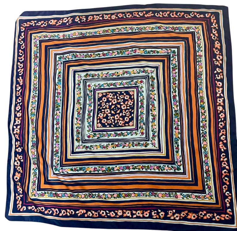 May include: Square silk scarf with a repeating floral and geometric pattern. The scarf features a navy blue border, orange stripes, and a central square with a floral design in shades of pink, green, and yellow.