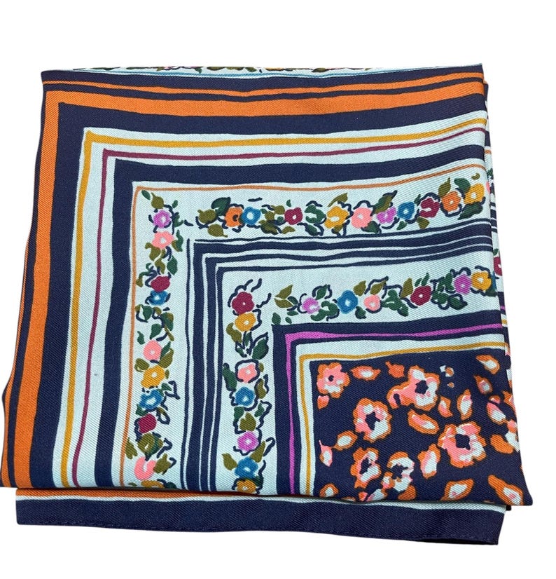 May include: Folded square scarf with a floral and striped pattern. The scarf features a navy blue border with orange and white stripes. The center has a white background with colorful flowers and a navy blue square.