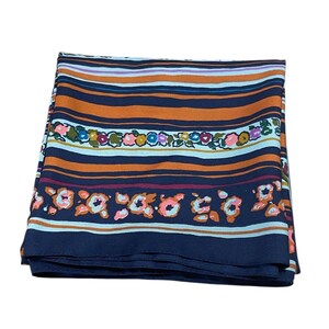 May include: Folded silk scarf with a striped pattern in shades of orange, navy blue, and light blue. The scarf features floral accents in pink, green, and orange. The scarf is folded and appears to be made of a lightweight material.