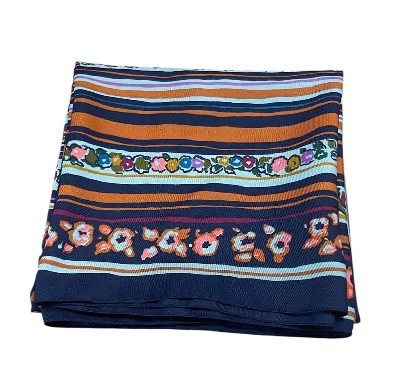 May include: Folded silk scarf with a striped pattern in shades of orange, navy blue, and light blue. The scarf features floral accents in pink, green, and orange. The scarf is folded and appears to be made of a lightweight material.