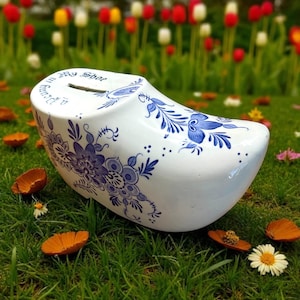 May include: White wooden shoe-shaped bank with blue floral designs. The bank has a slot for coins and the text "Put in my shoe" is visible. The bank is sitting on green grass with flowers and petals scattered around.